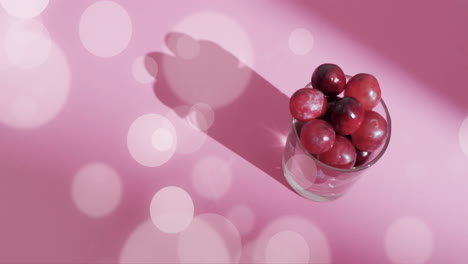 Composition-of-spots-of-light-over-glass-with-red-grapes-on-pink-background
