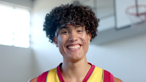 Young-biracial-man-with-curly-hair-smiles-brightly-in-a-basketball-court-setting