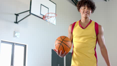 Young-biracial-man-with-curly-hair-holds-a-basketball-in-a-gym