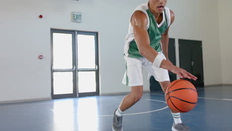 Young-biracial-man-in-a-green-jersey-dribbles-a-basketball-in-a-gym
