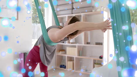 Animation-Von-Blauen-Und-Weißen-Lichtpunkten-über-Einer-Kaukasischen-Frau,-Die-Aerial-Yoga-Praktiziert
