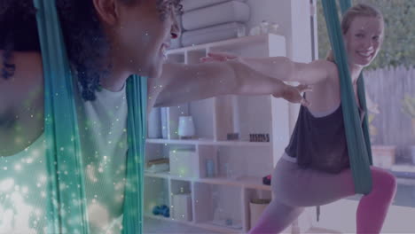 Animation-of-glowing-green-particles-over-happy-diverse-teenage-girls-practicing-aerial-yoga