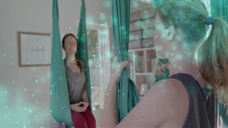 Animation-of-glowing-green-particles-over-happy-caucasian-female-aerial-yoga-teacher-and-student