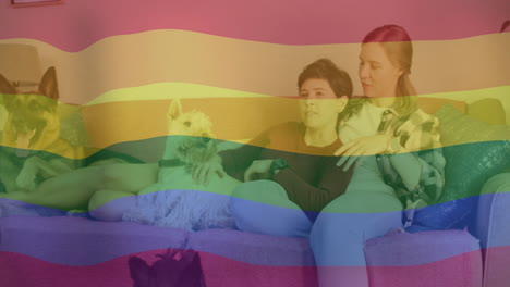 Animation-of-pride-rainbow-flag-over-happy-caucasian-lesbian-couple-relaxing-on-couch-with-pet-dogs