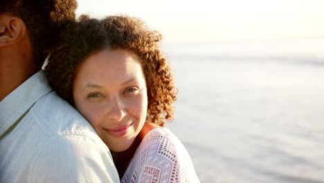 Biracial-couple-embraces,-woman-with-curly-hair-smiling-at-the-beach-during-sunset-with-copy-space