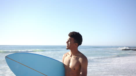A-young-biracial-man-holds-a-blue-surfboard-at-the-beach-with-copy-space