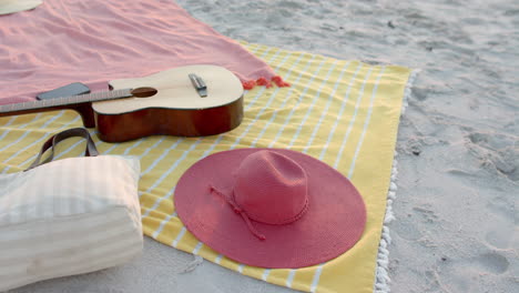 A-guitar-and-a-pink-hat-rest-on-a-beach-blanket,-with-copy-space