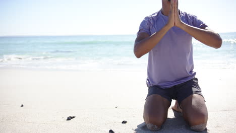 A-young-biracial-man-is-meditating-on-the-beach,-hands-pressed-together-in-a-prayer-pose,-with-copy-
