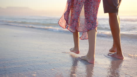 Biracial-couple-walks-barefoot-on-the-beach-at-sunset,-woman-wearing-a-patterned-dress,-with-copy-sp