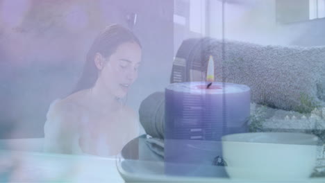 Animation-of-scented-candle-and-towels-over-caucasian-woman-in-bath-splashing-face