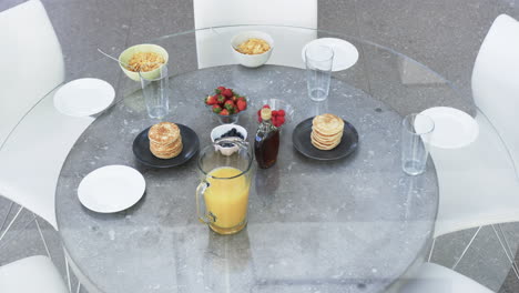 A-breakfast-spread-is-laid-out-on-a-glass-table-with-pancakes,-fruits,-and-juice