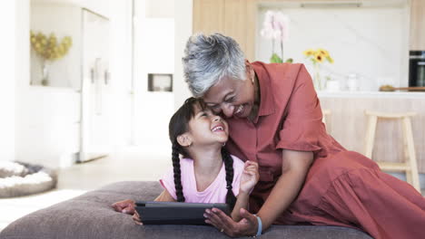 Biracial-grandmother-laughs-with-a-biracial-granddaughter-holding-a-tablet,-with-copy-space