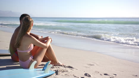 Young-Caucasian-woman-sits-on-a-surfboard-at-the-beach,-smiling-towards-the-camera-with-copy-space