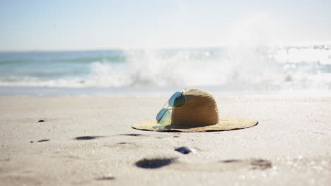 A-straw-hat-and-sunglasses-rest-on-a-sandy-beach,-with-the-ocean-in-the-background,-with-copy-space