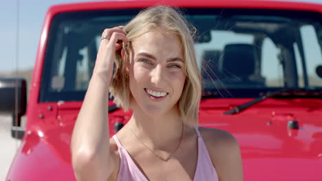 Young-Caucasian-woman-smiles-brightly-in-front-of-a-red-vehicle-on-a-road-trip