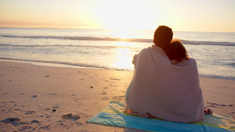 Biracial-couple-wrapped-in-a-blanket,-watching-a-beach-sunset-with-copy-space