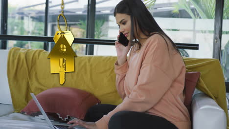 Animation-of-gold-house-key-and-key-fob-over-biracial-woman-using-laptop-on-sofa-at-home