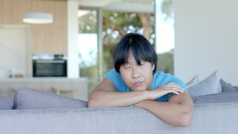 Thoughtful-teenage-Asian-boy-rests-his-chin-on-his-arms-atop-a-couch-at-home