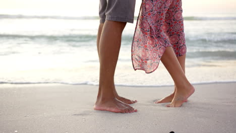 Barefoot-biracial-couple-stands-on-a-sandy-beach-at-sunset