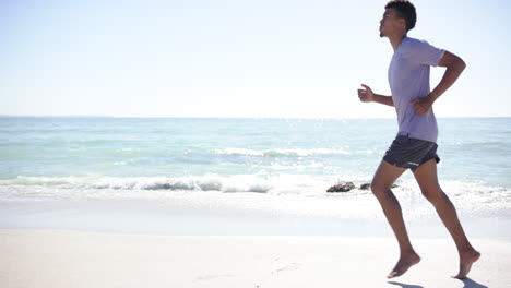 A-young-biracial-man-jogs-along-a-sunny-beach-with-copy-space,-his-athletic-form-accentuated-by-casu