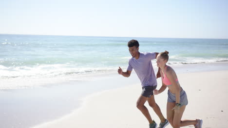 A-young-biracial-man-and-a-young-Caucasian-woman-are-jogging-on-a-sunny-beach-with-copy-space