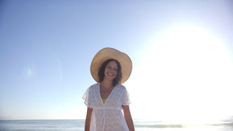 A-young-biracial-woman-stands-on-the-beach,-wearing-a-wide-brimmed-hat-and-a-white-cover-up