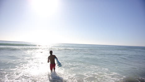 A-young-biracial-man-enters-the-sunlit-ocean-with-a-surfboard,-with-copy-space