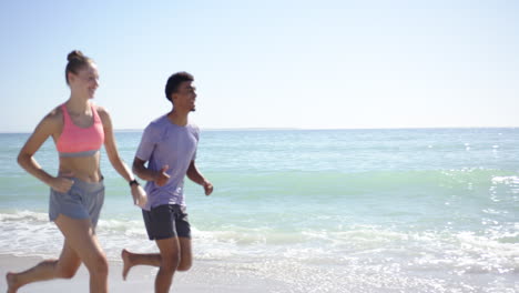 A-young-Caucasian-woman-and-a-biracial-man,-dressed-in-athletic-wear,-are-jogging-along-the-beach