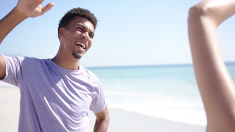 A-young-biracial-man-with-curly-hair,-wearing-a-purple-shirt,-smiles-joyfully-on-a-sunny-beach
