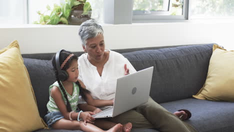 Biracial-grandmother-and-granddaughter-sit-on-a-couch,-engaged-with-a-laptop