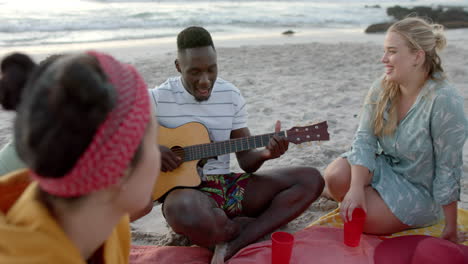 Young-African-American-man-plays-guitar-on-the-beach-at-a-party,-surrounded-by-friends