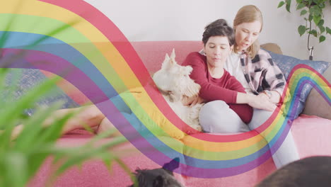 Animation-of-pride-rainbow-band-over-happy-caucasian-lesbian-couple-relaxing-on-couch-with-pet-dog