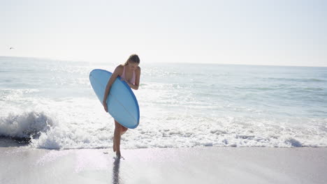 Young-Caucasian-woman-holds-a-blue-surfboard-on-a-sunny-beach-with-copy-space