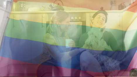 Animation-of-pride-rainbow-flag-over-happy-caucasian-lesbian-couple-sitting-with-pet-dog-in-kitchen