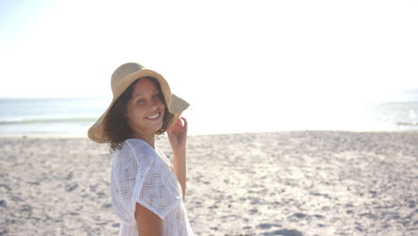 Young-biracial-woman-smiles-on-a-sunny-beach,-wearing-a-white-dress-and-a-straw-hat,-with-copy-space