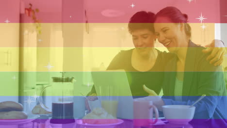 Animation-of-pride-text,-rainbow-stripes-over-happy-caucasian-lesbian-couple-using-tablet-at-table