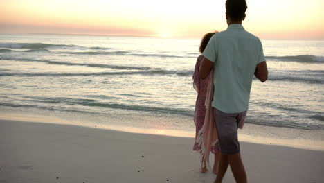 Biracial-couple-stands-close,-wrapped-in-a-pink-blanket,-watching-a-beach-sunset-with-copy-space