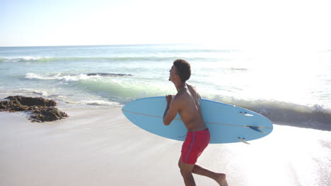Young-biracial-man-carries-a-light-blue-surfboard-on-a-sunny-beach
