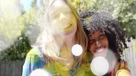 Animation-of-yellow-and-white-light-spots-over-two-happy-diverse-teenage-girls-embracing-in-garden