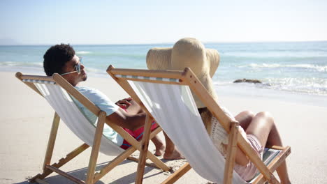 Biracial-man-and-young-Caucasian-woman-relax-on-beach-chairs-facing-the-sea