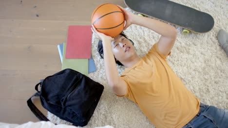 Asiatischer-Teenager-Liegt-Zu-Hause-Auf-Einem-Teppich-Und-Wirft-Einen-Basketball