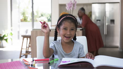 Biracial-girl-with-a-headband-smiles-while-doing-homework,-a-woman-in-the-background