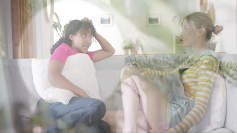 Animation-of-sunlight-and-plants-over-two-happy-diverse-teenage-girls-talking-on-couch