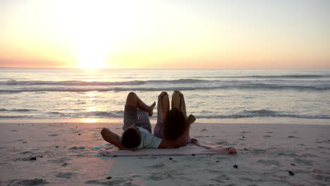 Biracial-couple-relaxes-on-a-beach-at-sunset,-lying-on-a-pink-towel