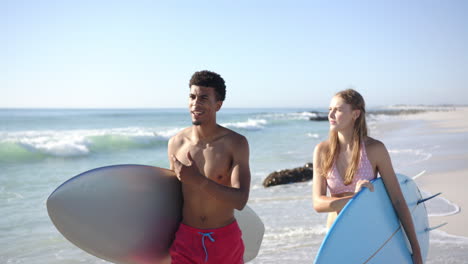 Young-biracial-man-and-Caucasian-woman-hold-surfboards-on-a-sunny-beach