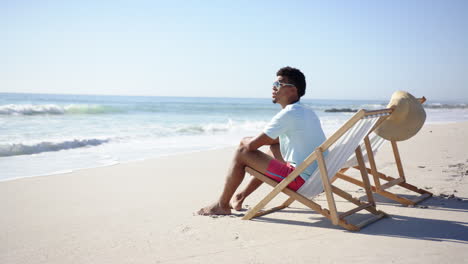 Young-biracial-man-relaxes-on-a-beach-chair-by-the-sea,-with-copy-space