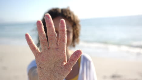 A-sandy-hand-is-raised-towards-the-camera,-obscuring-a-person's-face-at-the-beach