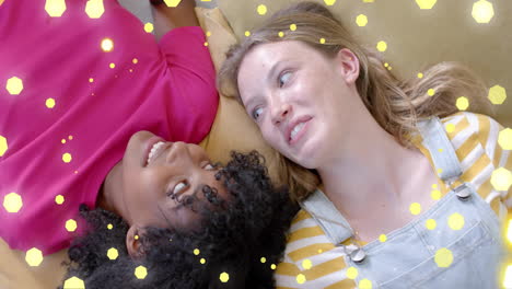 Animation-of-flashing-yellow-light-spots-over-two-happy-diverse-teenage-girls-lying-on-couch-talking
