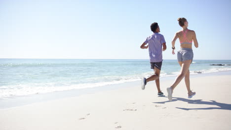 Young-biracial-man-and-Caucasian-woman-are-jogging-on-a-sunny-beach-with-copy-space