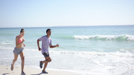 Young-Caucasian-woman-and-biracial-man-are-jogging-on-a-sunny-beach-with-copy-space